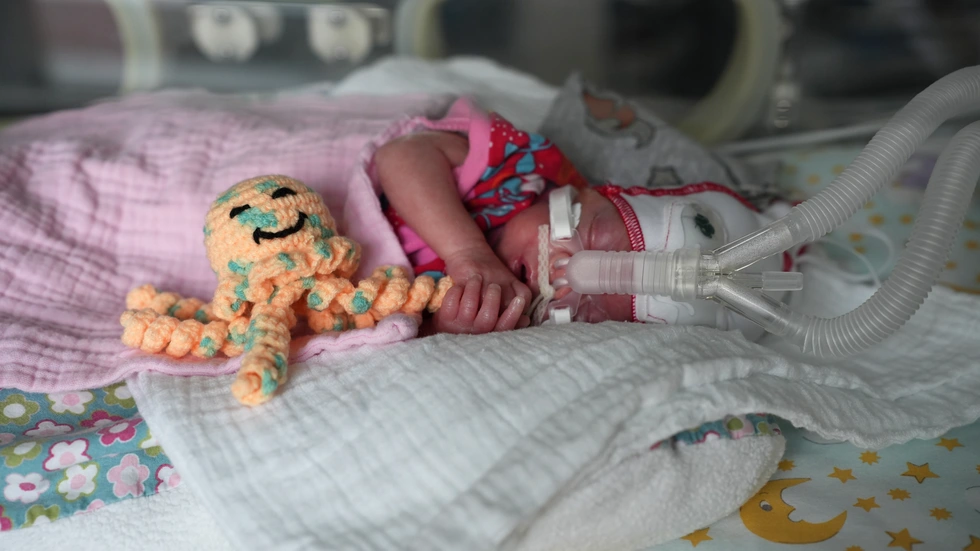 Emma liegt in ihrem Wärmebettchen, umgeben von Schläuchen, weichen Decken und gedämpftem Licht. Neben ihrem Gesicht sitzt eine kleine gehäkelte Krake. 